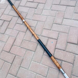 May include: A spear with a wooden shaft, black grip, and a silver blade. The spear is made of wood and metal. The shaft is a light brown color. The grip is black. The blade is silver. The spear is a traditional weapon.