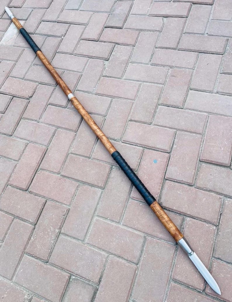 May include: A spear with a wooden shaft, black grip, and a silver blade. The spear is made of wood and metal. The shaft is a light brown color. The grip is black. The blade is silver. The spear is a traditional weapon.