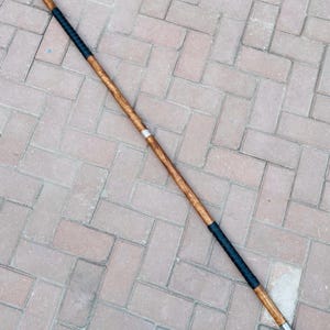 May include: A spear with a wooden shaft, metal spearheads, and black wrapped grips. The spear is brown and silver with a black grip. The spear is approximately 72 inches long.