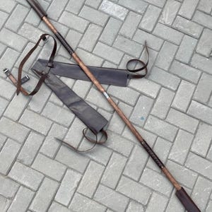 May include: A long, wooden spear with a dark brown blade and a leather sheath. The spear has a dark brown handle with a leather grip and a pointed tip. The sheath is also dark brown and has a leather strap.