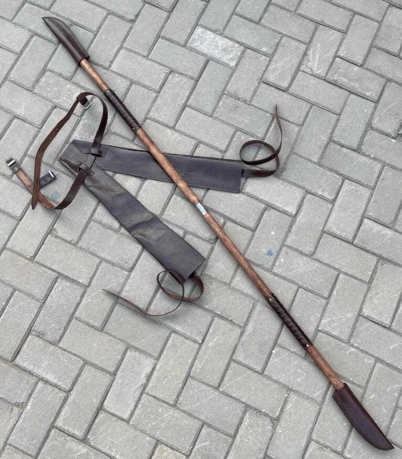 May include: A long, wooden spear with a dark brown blade and a leather sheath. The spear has a dark brown handle with a leather grip and a pointed tip. The sheath is also dark brown and has a leather strap.