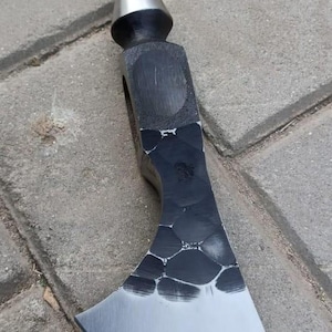 Op de afbeelding: Een handgesmede metalen bijl met een gepolijste zilverkleurige conische punt aan de bovenkant en een breed, gebogen blad. De bijlkop heeft een donkere, getextureerde afwerking met een opvallend gehamerd patroon dat overgaat in een rand.