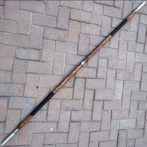 May include: A spear with a wooden shaft and a metal spearhead. The shaft is a light brown color with a black grip near the center. The spearhead is silver and tapers to a sharp point. The spear is lying on a brick surface.