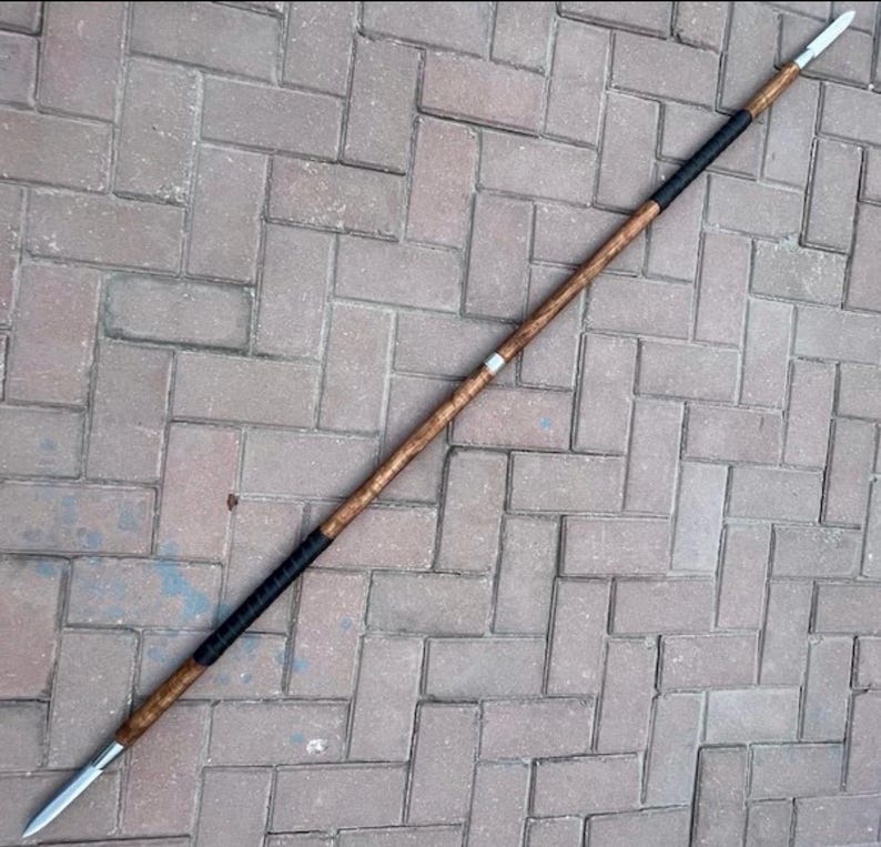 May include: A spear with a wooden shaft and a metal spearhead. The shaft is a light brown color with a black grip near the center. The spearhead is silver and tapers to a sharp point. The spear is lying on a brick surface.