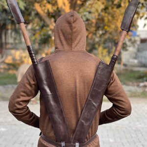 May include: A brown leather scabbard designed to hold two large blades, worn on the back. The blades have wooden handles and are sheathed in brown leather. The person is wearing a brown hooded sweatshirt and matching pants. The scabbard is secured with a belt.