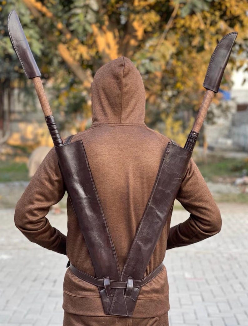May include: A brown leather scabbard designed to hold two large blades, worn on the back. The blades have wooden handles and are sheathed in brown leather. The person is wearing a brown hooded sweatshirt and matching pants. The scabbard is secured with a belt.