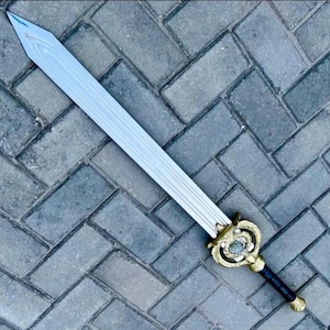 Puede incluir: Una espada larga y plateada con una empuñadura dorada decorativa y un mango negro. La hoja tiene un patrón texturizado. La empuñadura presenta un diseño ornamentado con una joya central. La espada se muestra sobre un fondo de ladrillos grises.