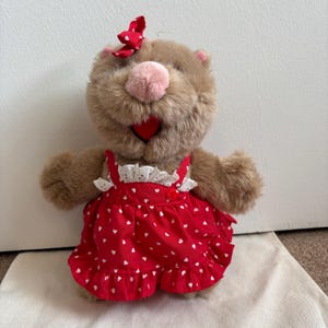 May include: A plush brown bear wearing a red dress with white heart patterns and a matching bow. The bear has a pink nose and a red heart-shaped mouth. The dress has white lace trim.