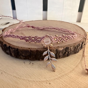 Könnte beinhalten: Ein altrosa Makramee-Halsband mit einer zentralen, durchscheinenden weißen Perle und einem silberfarbenen Blatt-Anhänger. Das Band ist mit kleinen Metallperlen verziert und verfügt über verstellbare Kordelenden mit kleinen runden Perlen.
