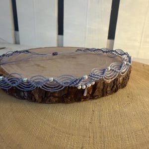 Könnte beinhalten: Eine hellblaue Makramee-Halskette mit komplizierten geknoteten Kordelmustern und kleinen, runden, metallisch silbernen Perlen. Das Design umfasst einen mittigen, tropfenförmigen silbernen Anhänger, der an der Unterkante der gewebten Struktur hängt.