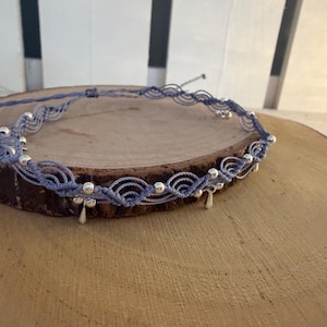 Op de afbeelding: Een lichtblauwe macramé choker ketting met een reeks herhalende golfvormige knopen. Het ontwerp is geaccentueerd met kleine, ronde, gepolijste zilverkleurige kralen en verschillende bungelende zilverkleurige traanvormige bedels.