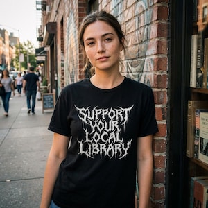 May include: Black t-shirt with the text "SUPPORT YOUR LOCAL LIBRARY" in a white, gothic-style font. The shirt is worn by a person standing in front of a brick wall and a bookstore.