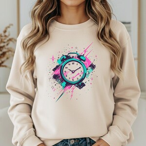 May include: A sand-colored crewneck sweatshirt featuring a graphic print of an alarm clock in shades of teal, pink, and black. The design includes abstract geometric shapes, starbursts, and energetic brushstroke patterns surrounding the clock face.