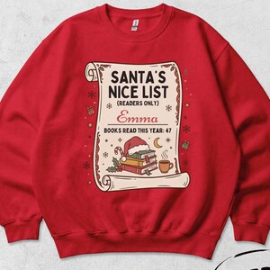 May include: A red crewneck sweatshirt featuring a graphic of a scroll titled SANTA'S NICE LIST (READERS ONLY). The design includes the name Emma, the text BOOKS READ THIS YEAR: 47, a stack of books with a Santa hat, a crescent moon, and a coffee mug.