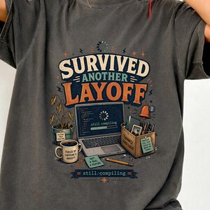 May include: Dark grey t-shirt with the text "Survived Another Layoff" in orange and white. The graphic includes a laptop, coffee mug, and a box of office supplies. The phrase "still compiling" is also present.
