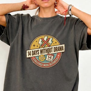 May include: Dark gray t-shirt with a round graphic that reads "14 Days Without Drama." The graphic includes lightning bolts, stars, and the words "Challenge Mode Activated."