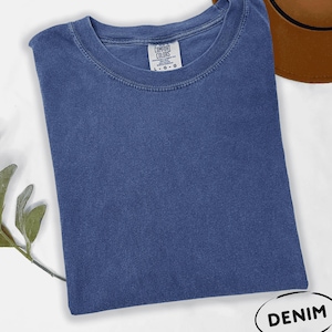 May include: A solid blue short-sleeve t-shirt with a crew neckline. The inner neck label reads Comfort Colors and indicates a size large. The fabric has a slightly faded, washed appearance. A small white oval graphic in the corner says DENIM.