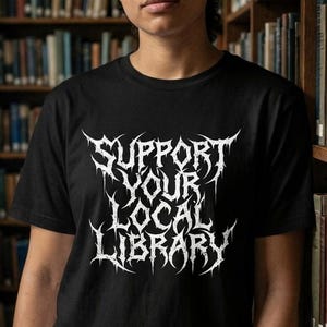 May include: Black t-shirt with the white text "SUPPORT YOUR LOCAL LIBRARY" in a stylized font. The person is standing in a library with bookshelves filled with books.