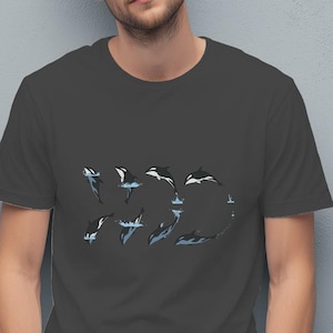 May include: Dark gray t-shirt featuring a graphic of orcas in a circular pattern. The orcas are depicted in shades of white, blue, and black, creating a dynamic and eye-catching design. The shirt is made of a soft material.
