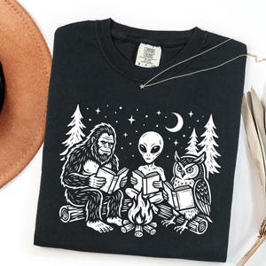May include: A black crewneck t-shirt featuring a white graphic print of a Bigfoot, an alien, and an owl sitting on logs around a campfire reading books under a crescent moon and stars. The inner neck label displays the Comfort Colors brand logo.