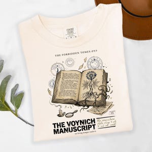 May include: A cream-colored t-shirt featuring a graphic of an open book with a plant illustration, celestial circles, a candle, and a magnifying glass. The text reads THE FORBIDDEN TOMES, THE VOYNICH MANUSCRIPT, and no living tongue reads it.
