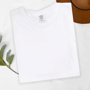 May include: A folded white t-shirt with a crew neck, featuring a tag that reads "COMFORT COLORS". A brown hat is partially visible in the upper right corner. A sprig of green leaves is in the lower left corner. The shirt is made of a soft material.