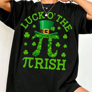 May include: Black t-shirt with a green graphic design. The design features the text "LUCK O' THE IRISH" with a green leprechaun hat and a pi symbol made of shamrocks. Several shamrocks are scattered around the design.