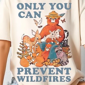 以下が含まれることがあります： クリーム色のTシャツ。スモーキーベアと森の動物たちのグラフィックが特徴です。「ONLY YOU CAN PREVENT WILDFIRES」の文字が青色でデザインされています。イラストはヴィンテージ風の漫画スタイルです。
