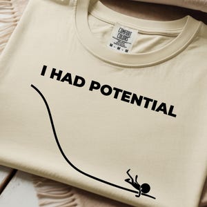 May include: A cream-coloured t-shirt featuring the text "I HAD POTENTIAL" in bold black lettering. A black stick figure is shown at the end of a curved black line, creating a humorous image. The shirt has a classic crew neck and a Comfort Colors label.