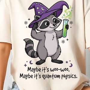 Op de afbeelding: Crèmekleurig T-shirt met een cartoon wasbeer met een paarse tovenaars hoed met sterren en manen. De wasbeer houdt een reageerbuis met groene vloeistof vast. De tekst op het shirt luidt: "Maybe it's woo-woo. Maybe it's quantum physics."