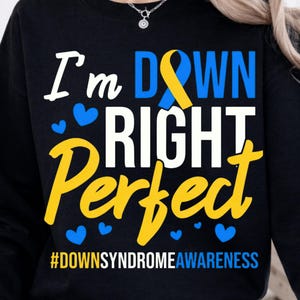 May include: Black sweatshirt with the text "I'm DOWN RIGHT Perfect" in white, yellow, and blue. The design includes a blue and yellow ribbon and blue heart shapes. The hashtag #DOWNSYNDROMEAWARENESS is at the bottom.