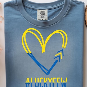May include: A blue t-shirt featuring a yellow and blue heart and arrow design, with the text "#LUCKYFEW" in blue. The shirt is made by Comfort Colors.