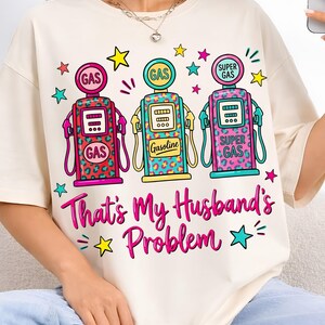 Könnte beinhalten: Cremefarbenes T-Shirt mit einer Grafik von drei Retro-Zapfsäulen in Pink, Türkis und Gelb mit Leopardenmuster-Akzenten. Der Text "That's My Husband's Problem" ist in Pink und Türkis geschrieben. Sterne umgeben die Grafik.