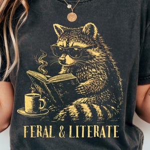 May include: Dark gray t-shirt featuring a raccoon wearing glasses and reading a book, with a cup of coffee on a saucer. The text "FERAL & LITERATE" is printed below the illustration in a gold color.
