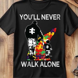 May include: Black t-shirt with the words "YOU'LL NEVER WALK ALONE" in white. The shirt features a graphic of a parent and child holding hands, with puzzle pieces and a puzzle piece symbol.