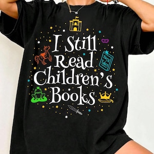 Puede incluir: Camiseta negra con el texto blanco "I Still Read Children's Books". El diseño incluye un castillo, un caballo, una rana, una corona, un cuchillo y un libro con "Fairy Tales". El diseño está rodeado de estrellas.