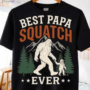 Può includere: T-shirt nera con una grafica raffigurante un essere simile a Bigfoot e un bambino, con la scritta "BEST PAPA SQUATCH EVER". Il design include montagne e alberi in verde, marrone e bianco.