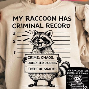 May include: Beige sweatshirt with the text "MY RACCOON HAS CRIMINAL RECORD" above a raccoon illustration. The raccoon holds a sign that reads "CRIME: CHAOS. DUMPSTER RAIDING. THEFT OF SNACKS." The design includes a height chart.