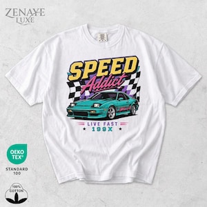 Puede incluir: Una camiseta blanca de manga corta con un gráfico de un coche deportivo color cerceta con faros escamoteables. El texto dice SPEED Addict, LIVE FAST y 199X. Detrás del coche hay un patrón de bandera a cuadros en blanco y negro con detalles en rosa y amarillo.