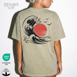 May include: A sage green short-sleeve t-shirt featuring a graphic on the back of a large black stylized ocean wave, a solid red sun, and three small black birds in flight. The text ZENAYE LUXE is printed in the upper left corner of the shirt.