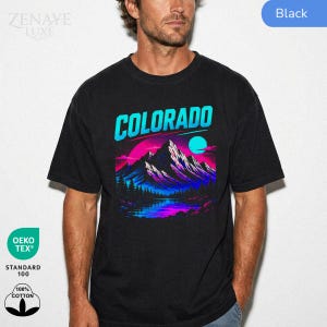 May include: A black short-sleeve t-shirt featuring a graphic print of a mountain range with a lake in the foreground. The design uses vibrant neon blue, purple, and pink colors with the word COLORADO printed in large cyan letters at the top.