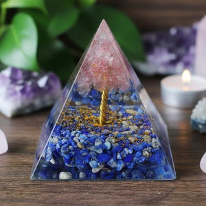 Pode incluir: Uma pirâmide de resina transparente com um design de árvore rosa e dourada. A base é preenchida com pedras azuis e brancas. Uma árvore dourada eleva-se das pedras, encimada por cristais rosa. A pirâmide está sobre uma superfície de madeira.