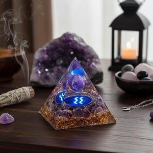 May include: A clear pyramid-shaped object with purple stones and gold flakes inside. The pyramid has blue lights and symbols. Other items on the table include a large amethyst, a candle in a black lantern, and a bowl of stones.