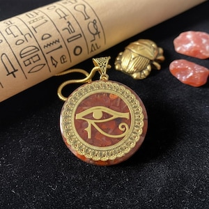 Puede incluir: Colgante dorado con el símbolo del Ojo de Horus sobre un fondo rojo. El colgante redondo tiene un borde decorativo y cuelga de una cadena dorada. También se ven un escarabajo dorado y dos piedras rojas.
