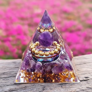 Peut inclure: Une pyramide transparente avec une sphère d'améthyste violette et des accents dorés. La pyramide contient des cristaux d'améthyste et des paillettes d'or. Un anneau de perles dorées entoure un motif circulaire. L'arrière-plan est rose flou.