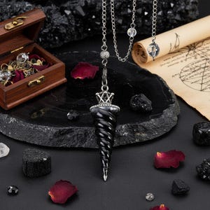 Puede incluir: Un péndulo negro en espiral con una cadena plateada y un diseño de corona. El péndulo se exhibe sobre una superficie oscura y texturizada con cristales dispersos, pétalos de rosa y una pequeña caja de madera.