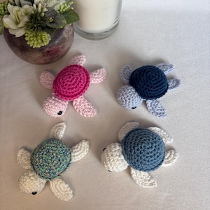 Pode incluir: Quatro pequenas figuras de tartarugas marinhas feitas de crochê. Uma tem uma carapaça rosa brilhante com membros rosa claro, uma tem uma carapaça azul escura com membros azul claro, uma tem uma carapaça multicolorida com membros creme.