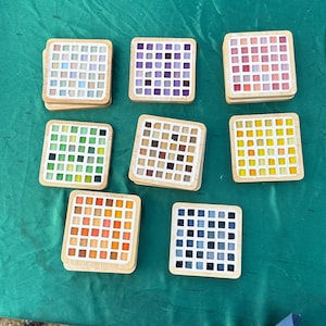 May include: Eight square coasters with light wood frames, each featuring a grid of small, colored glass tiles. The sets display various color palettes including white, purple, pink, green, brown, yellow, orange, and dark blue glass squares.
