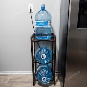 May include: A dark brown wooden water jug storage rack with three tiers. The top tier holds one large clear plastic water jug upright, while the middle and bottom tiers each hold one large clear plastic water jug positioned on its side.
