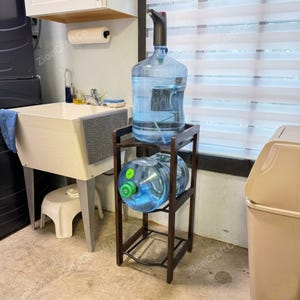 May include: A dark brown wooden water jug stand featuring two tiers designed to hold large plastic water bottles. The top tier supports an upright bottle with a black electric pump, while the bottom tier holds a second bottle lying horizontally.